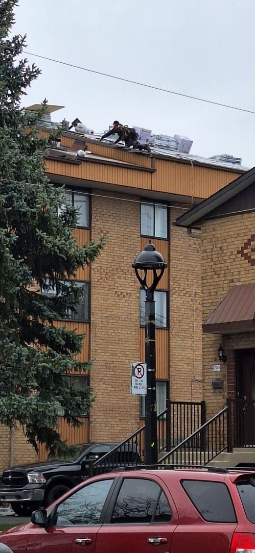 Un ouvrier vêtu de sombre travaille sur le toit d'un immeuble en briques de plusieurs étages, avec un lampadaire et des véhicules en contrebas.