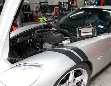 Silver sports car in a garage with hood open and engine exposed, black racing stripes visible.