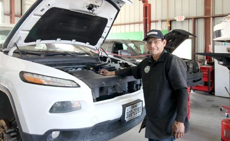 A mechanic in a shop stands next to a white SUV with its hood open, smiling at the camera.