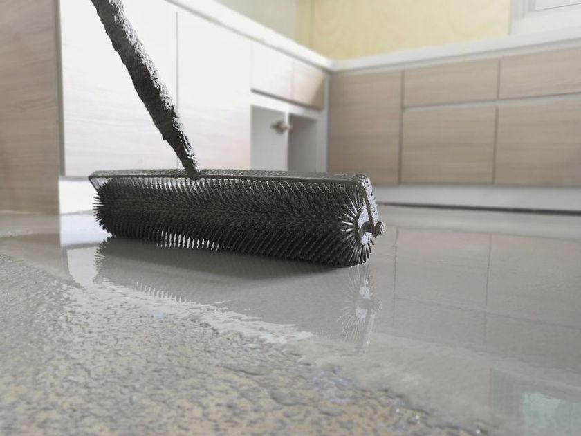 A person using a spiked roller on a wet, gray epoxy floor in a kitchen.