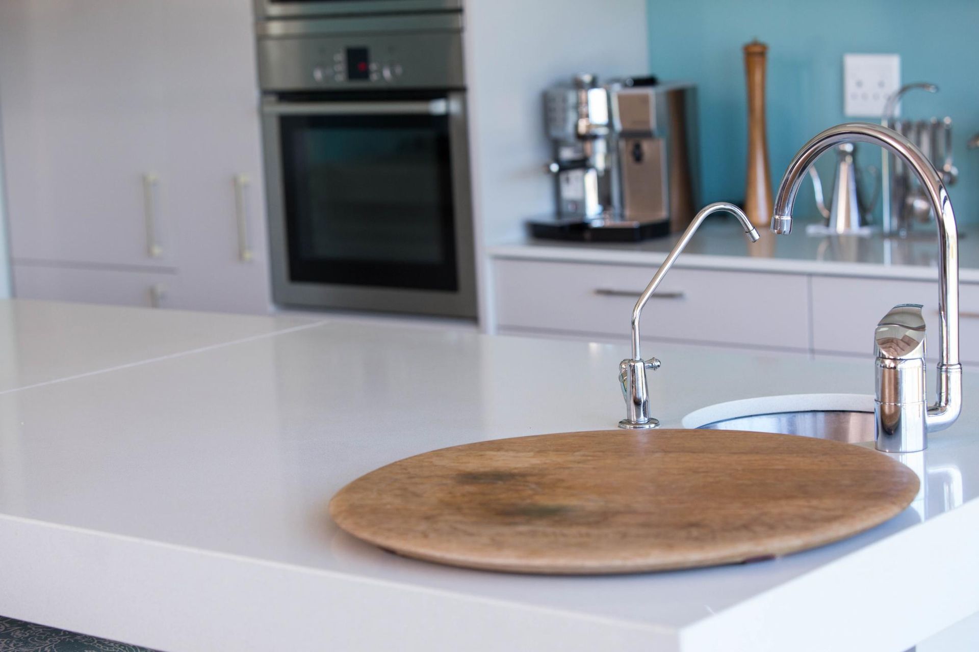 A white countertop with a cutting board and faucet in a kitchen. An oven and coffee machine are in the background.