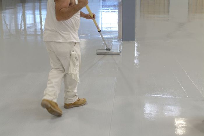 A person rolls glossy gray sealant onto a large floor, wearing white work clothes.