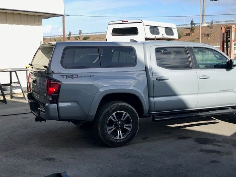 A gray Toyota Tacoma TRD Sport pickup truck with a camper shell parked on a paved lot.