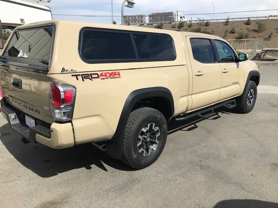 Tan Toyota Tacoma TRD 4x4 pickup truck with a matching camper shell parked in an asphalt lot.