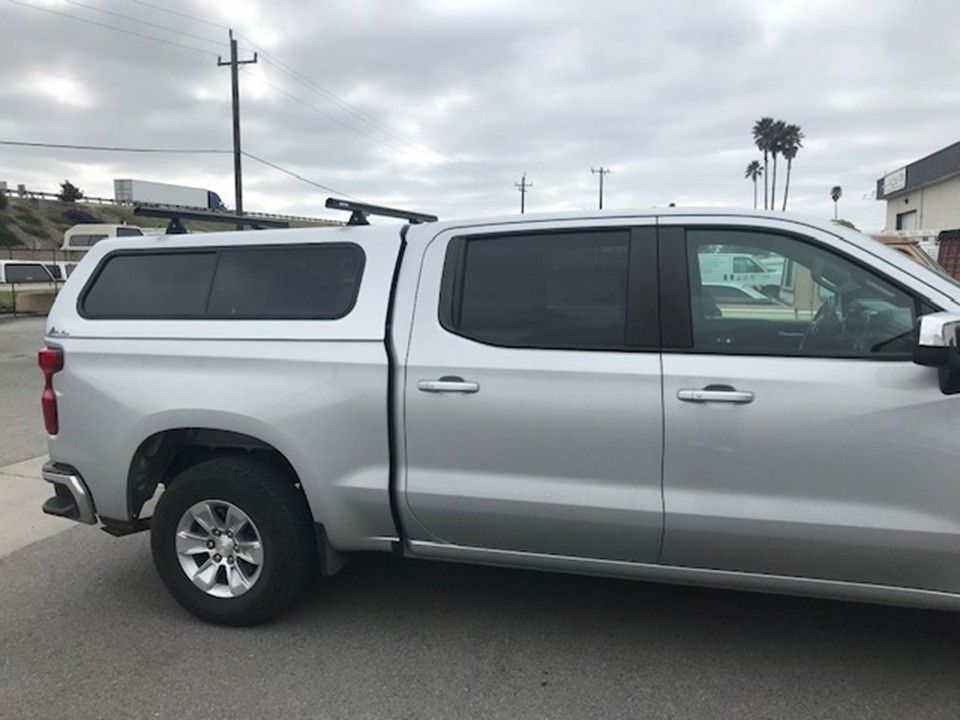A silver crew cab pickup truck with a matching canopy shell parked outdoors under a cloudy sky.