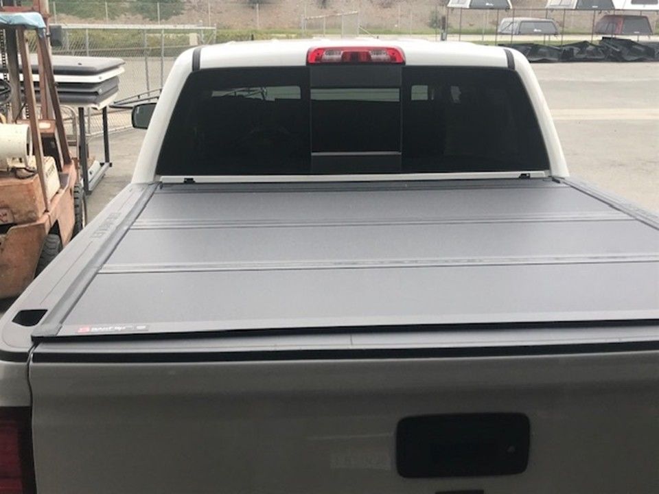 A white pickup truck with a black folding tonneau cover installed on the truck bed, parked in an outdoor lot.