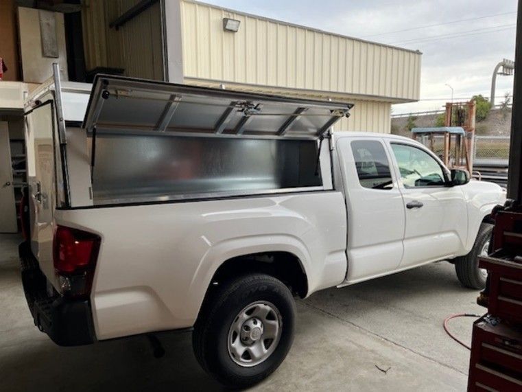A white pickup truck with an open metal utility box topper parked inside a workshop.
