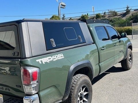 An olive green Toyota Tacoma TRD truck with a black camper shell parked outdoors on a sunny day.