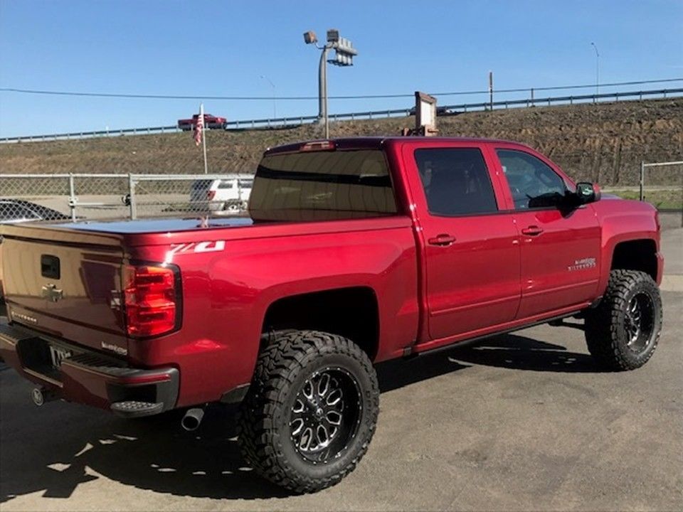 A red crew cab pickup truck with large off-road tires and a black bed cover parked on an asphalt lot.