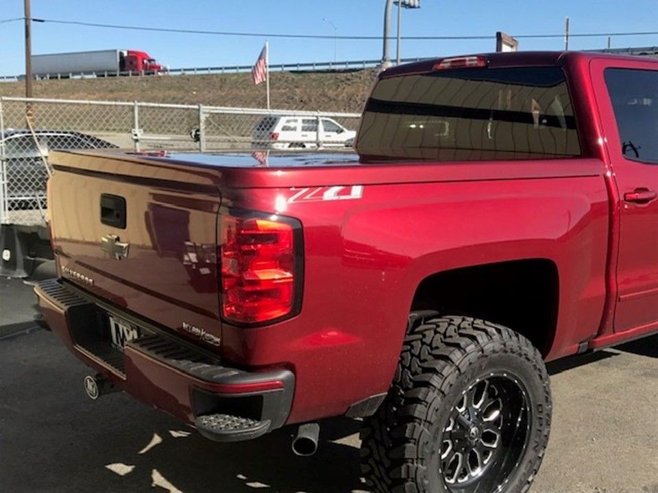 A red pickup truck with a bed cover parked on an asphalt lot with a highway and an American flag in the background.