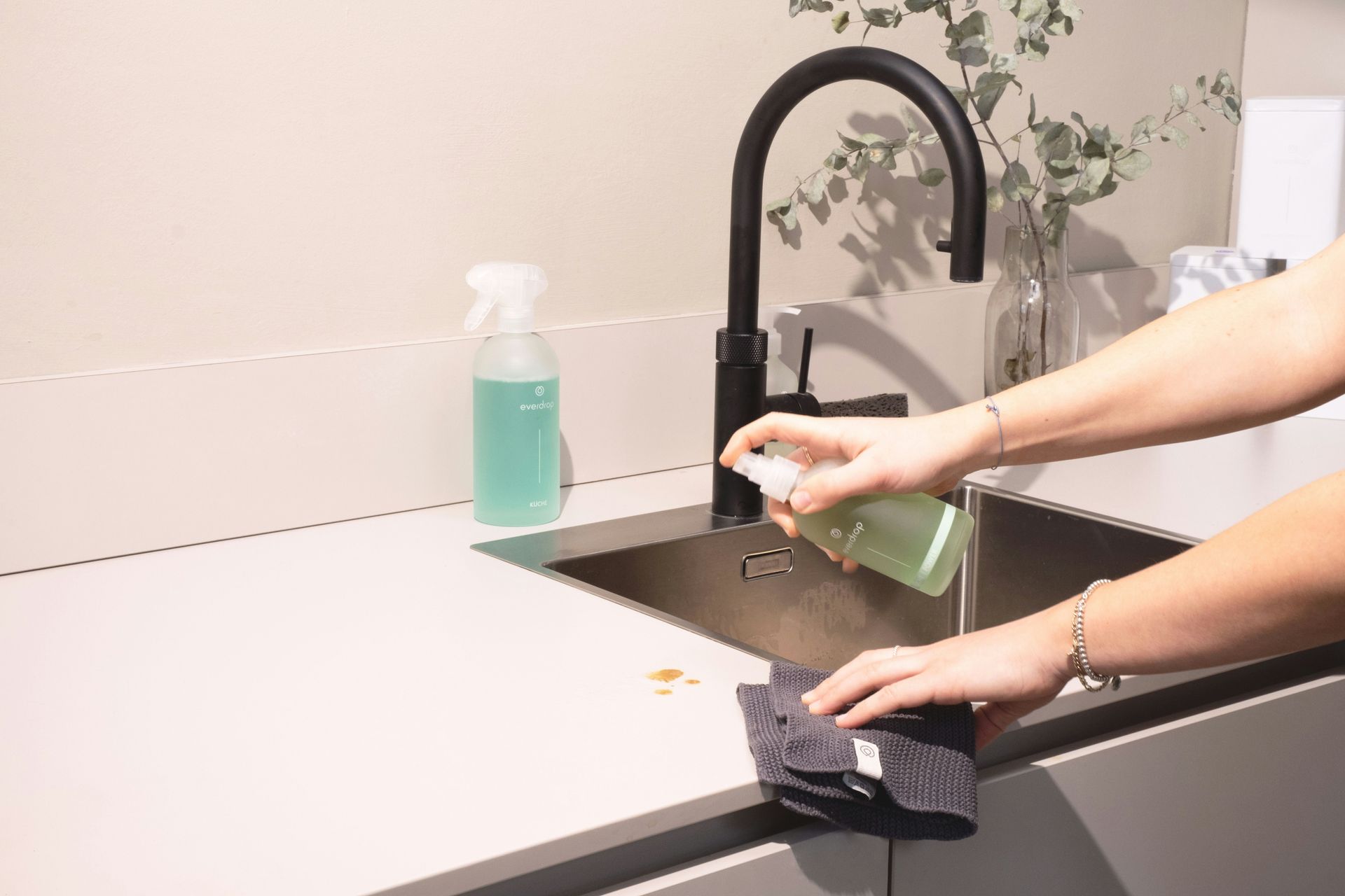 Person spraying and wiping a kitchen countertop with a cleaning product near a sink and faucet.