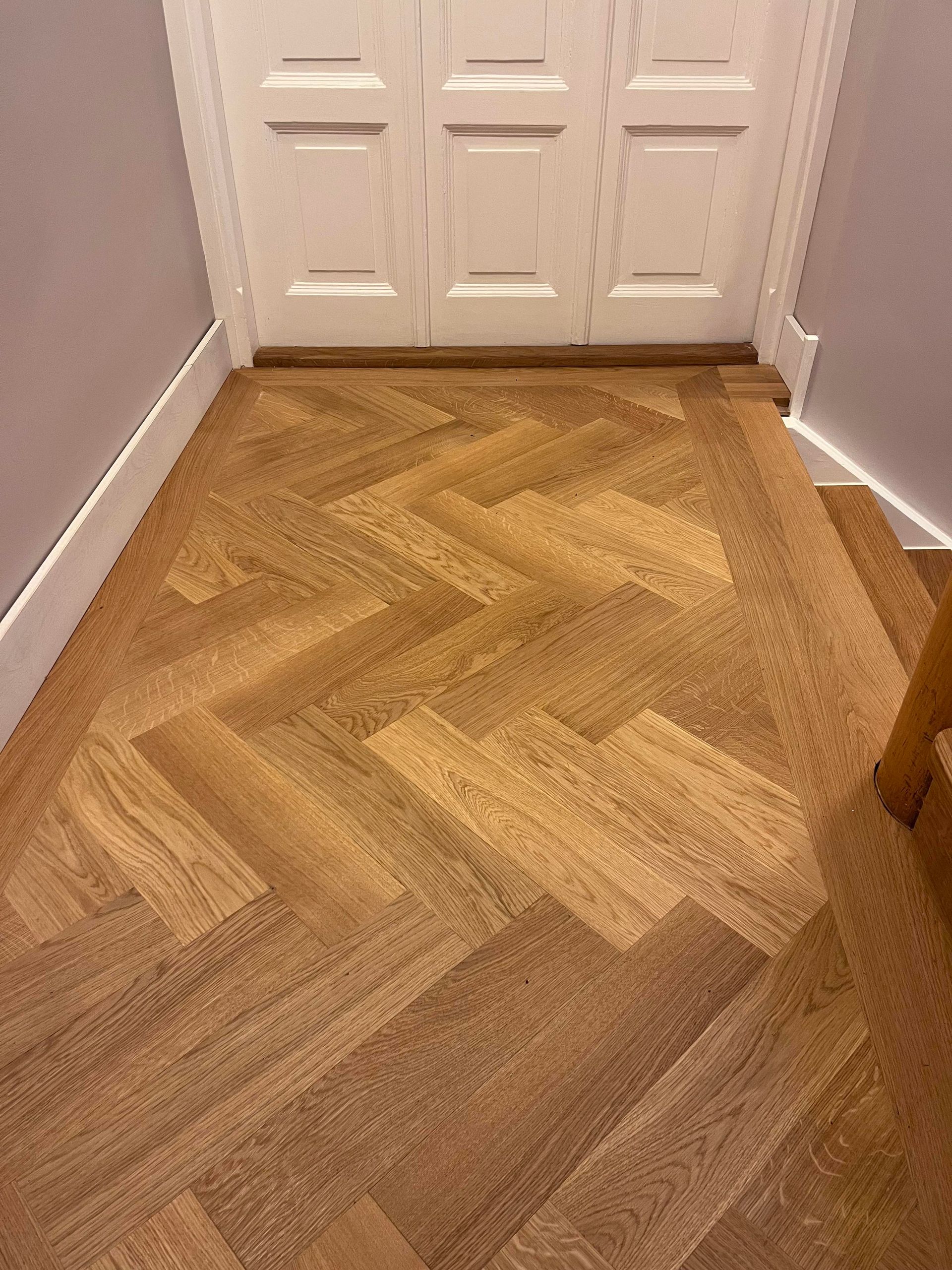 Ein Holzboden im Fischgrätmuster führt zu einer weißen Tür, rechts davon befindet sich eine Treppe und die Wände sind grau.