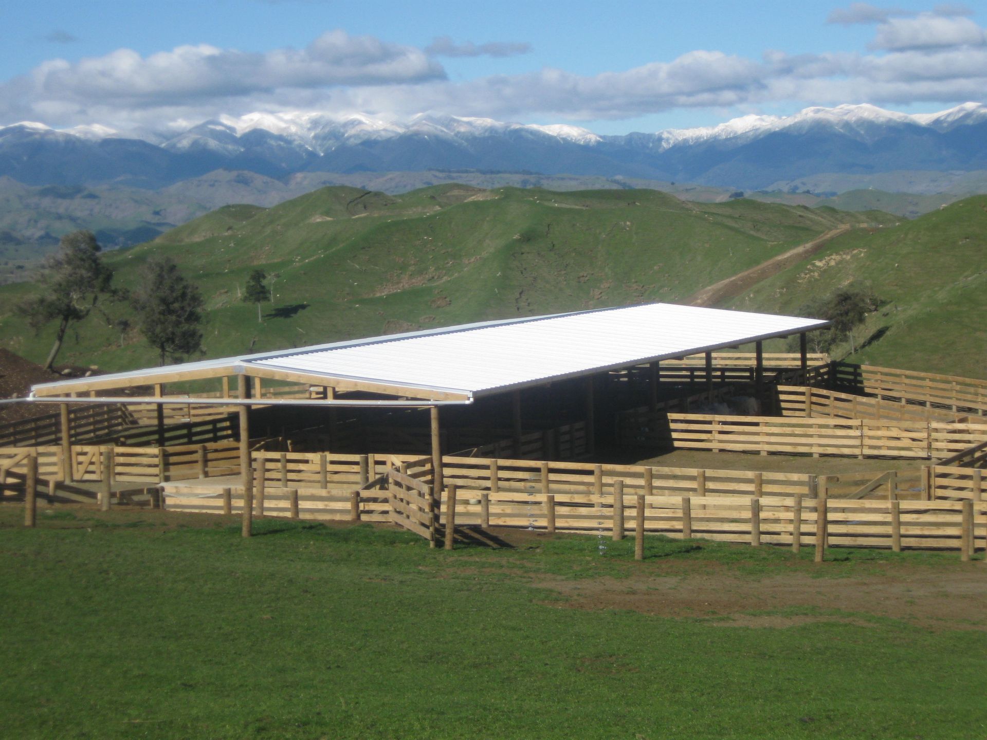 A large wooden cattle yard with a white roof, situated in a green grassy valley with snowy mountains in the background.