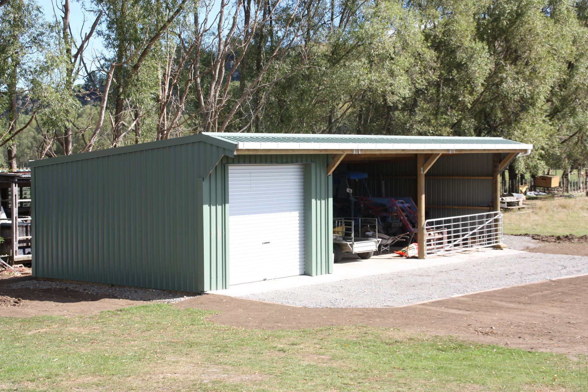 Green metal shed with a roller door and an open-sided carport, situated on a rural property with trees in the background.