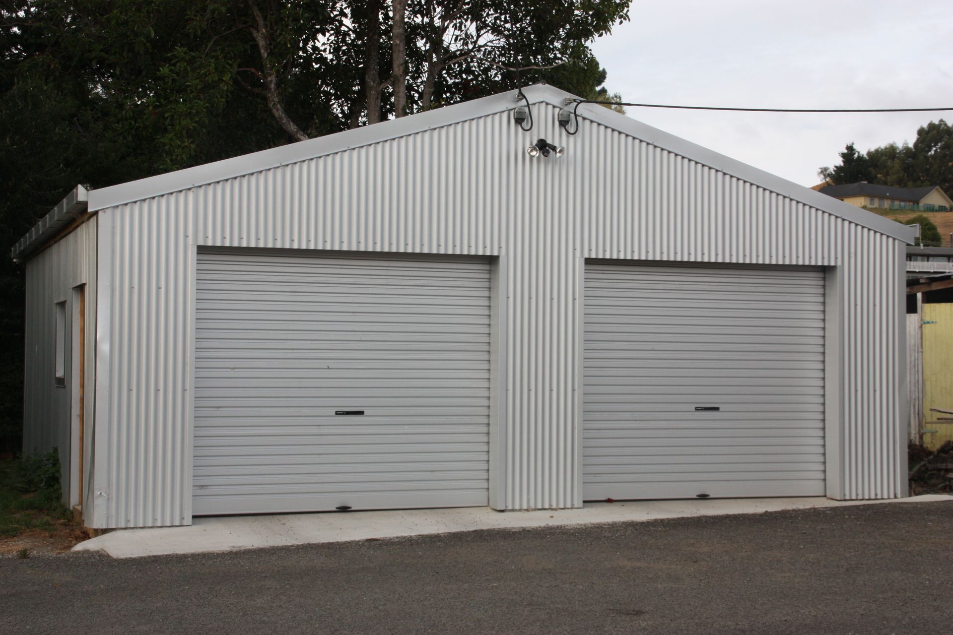 A detached double garage with a light gray corrugated metal exterior and two roll-up doors, set on a gravel lot.