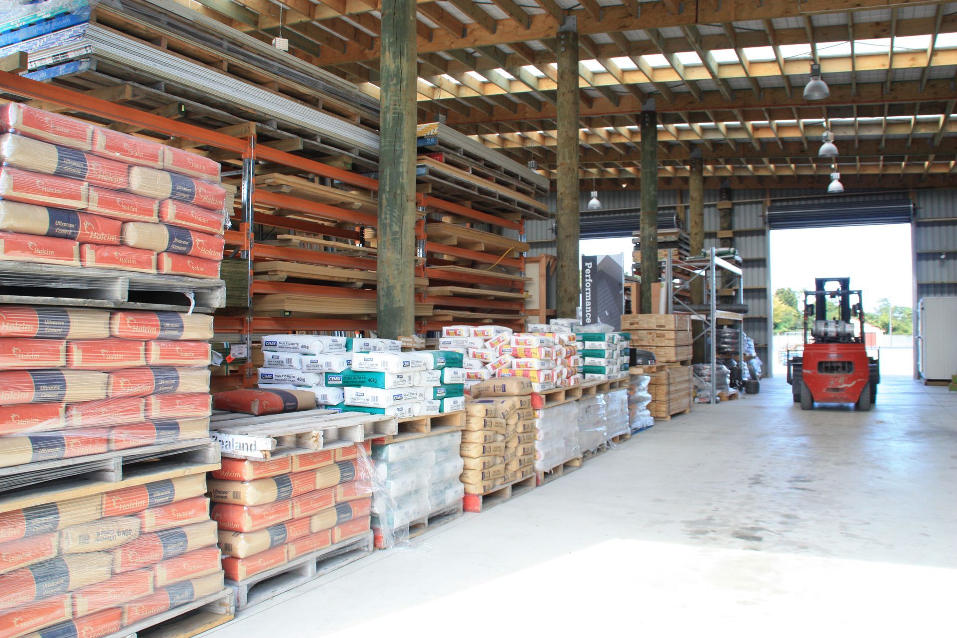 A forklift parked in a warehouse aisle filled with pallets of bagged construction materials and stacked lumber.