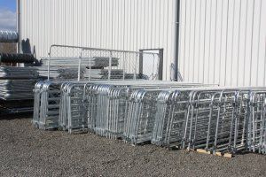 Stacks of galvanized steel livestock gates resting on a gravel lot against a white metal building.