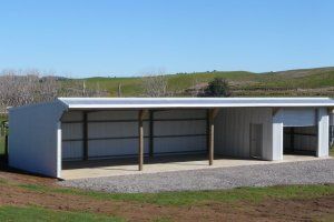 A white metal farm shed with an open-sided bay and an enclosed section, set on a gravel base in a grassy field.