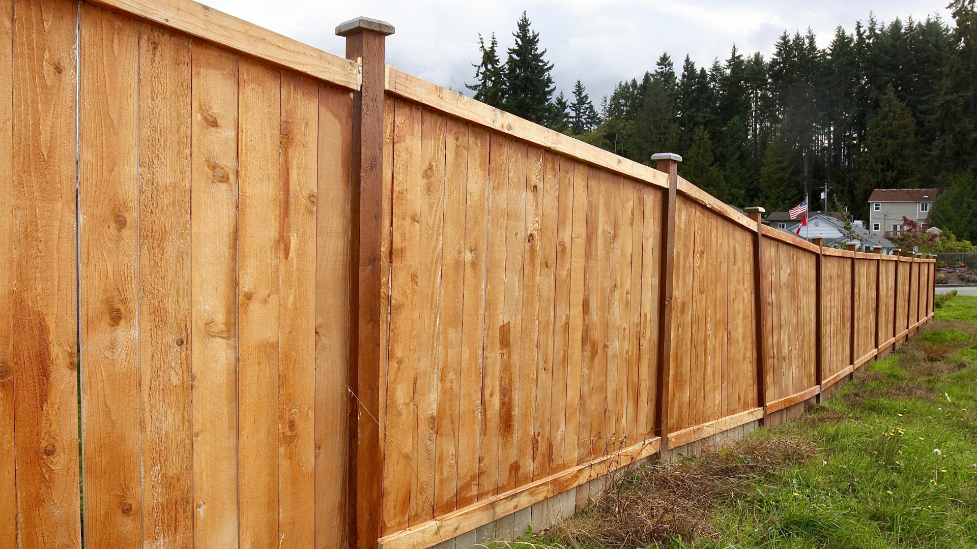 Wooden fence with vertical planks, brown stain, with posts and decorative caps, in front of greenery.