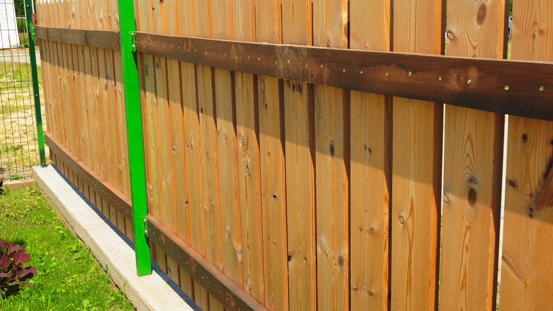 Wooden fence with brown horizontal beams, green post, and concrete base in a grassy yard.