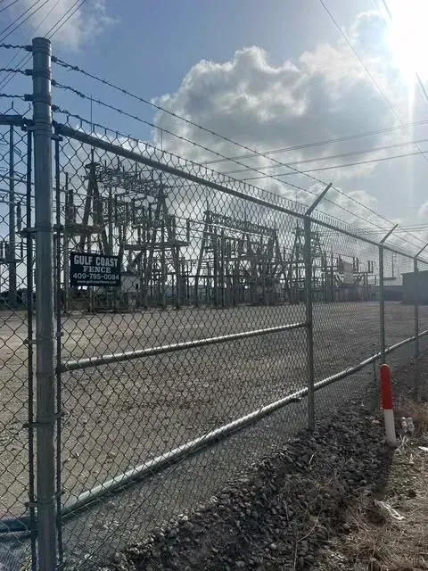 Chain-link fence topped with barbed wire encloses an electrical power substation under a cloudy sky.