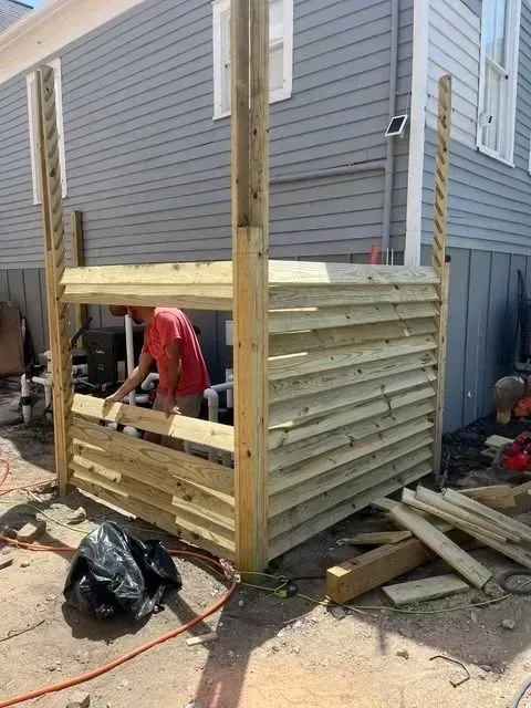 Construction of a wooden enclosure, likely for equipment. Person in red shirt works on the structure next to a gray house.