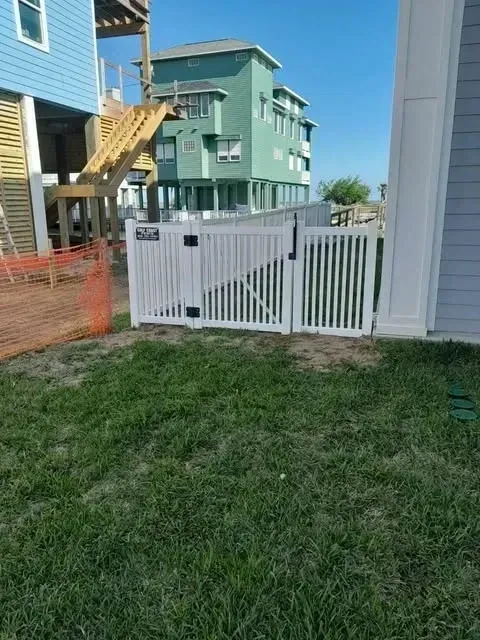 White picket fence with gate in front of a blue and green house, grass in the foreground.