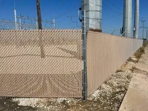 Chain-link fence with beige privacy slats. Gravel and a sidewalk are at the base, power lines are in the background.