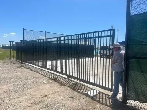 Black metal security gate, partially open, with a person in jeans and hard hat standing beside it. Sunny outdoor setting.