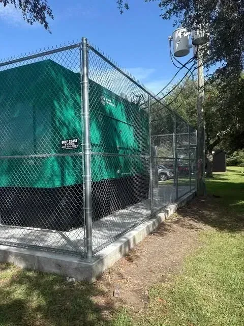 Generator inside a chain link fence, green cover, concrete base, and utility pole with transformer in a grassy area.