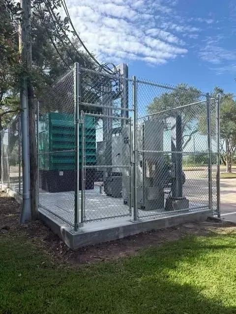 Fenced electrical equipment enclosure on a concrete pad with a utility pole nearby, under a blue sky.
