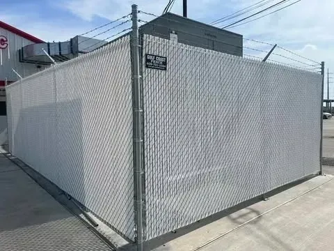White privacy slats in a chain-link fence enclose an industrial area. Barbed wire tops the fence.