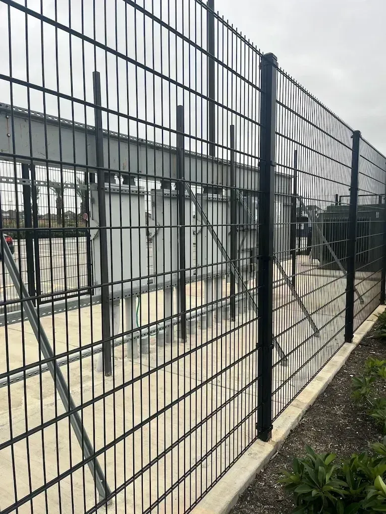 Black metal security fence surrounding an industrial area with equipment and concrete foundation.