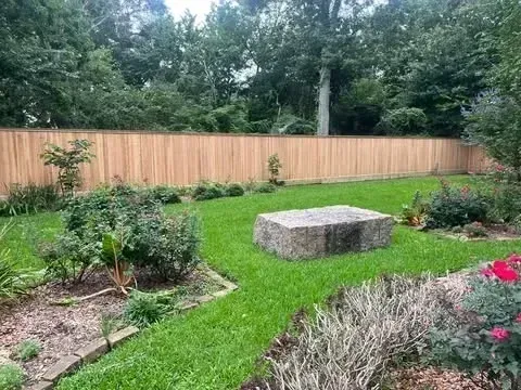 A wooden fence surrounds a green backyard with a rock centerpiece, lush plants, and trees in the background.