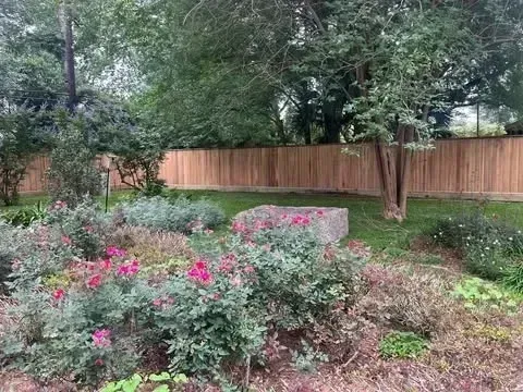 A flower garden with pink blooms, a stone block, and a wooden fence, under green trees.