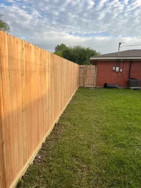 Wooden fence along a grassy backyard, with a gate and a brick house visible in the background.