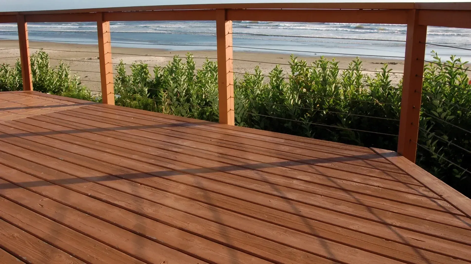 Wooden deck overlooking a sandy beach and ocean, with wooden railing and green shrubs.