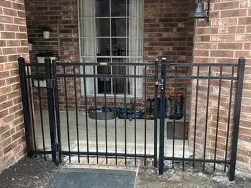 Black metal fence and gate on a brick patio, in front of a window.