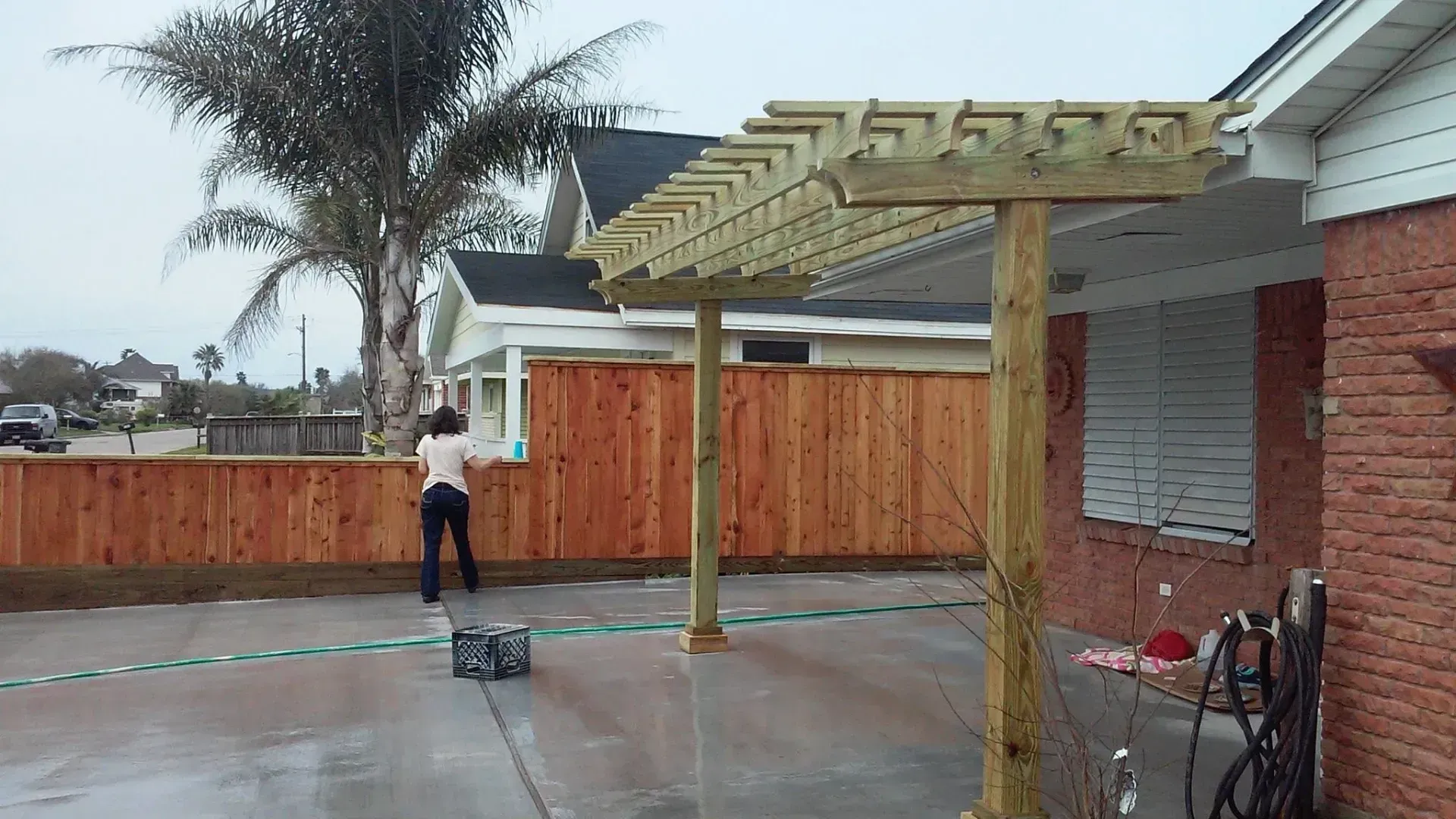 Woman cleaning a wet patio with a hose, near a wooden fence and arbor.