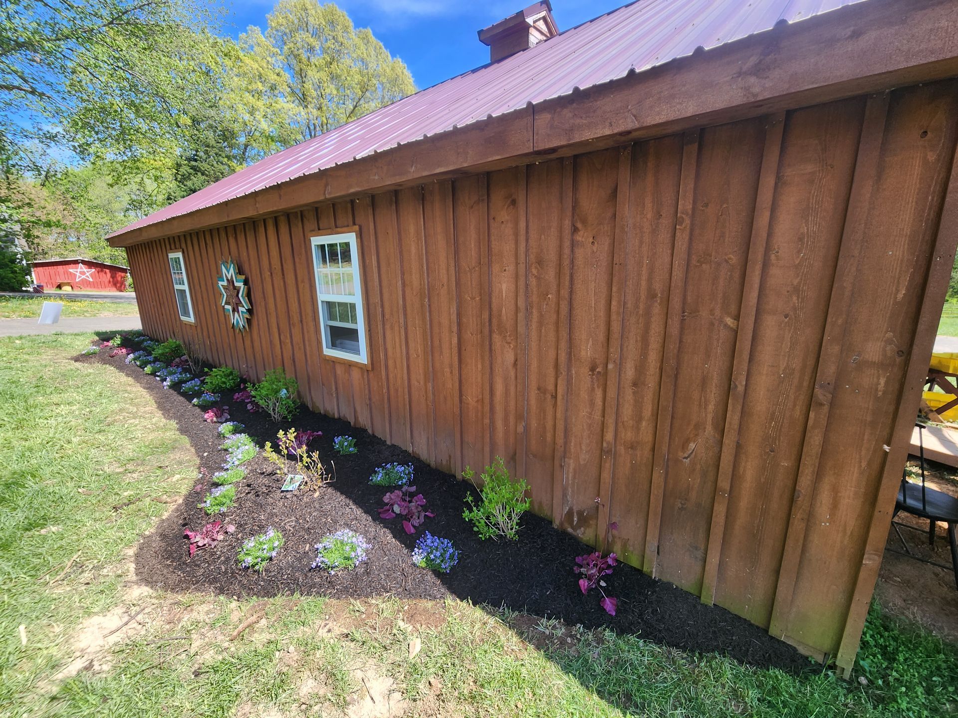Brown barn with a flower bed against it; green grass, blue sky, and trees.