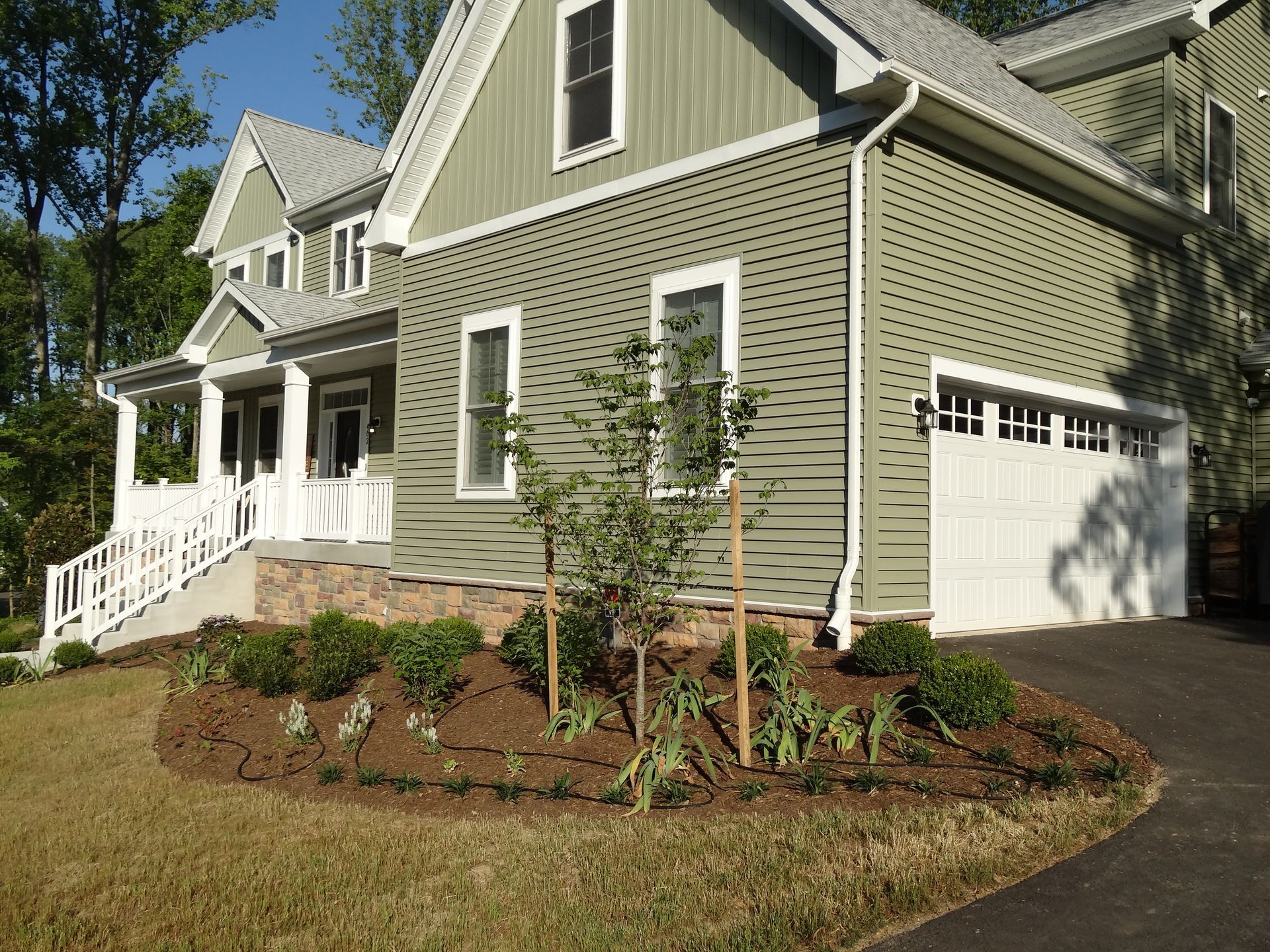 Two-story house with green siding, white trim, and a paved driveway. Landscaping with new trees and mulch in front.