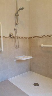 Doccia accessibile con sedile ribaltabile, maniglioni e soffione doccia portatile. Piastrelle beige e pavimento bianco.