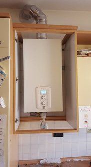 Una caldaia bianca installata all'interno di un mobiletto di legno in cucina. Un tubo di scarico argentato si collega alla parte superiore.