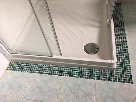Box doccia con base bianca e porte in vetro, circondato da finiture e pavimento in piastrelle a mosaico.