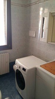 Bagno con lavatrice, piccola finestra, pareti e pavimento piastrellati e termosifone.