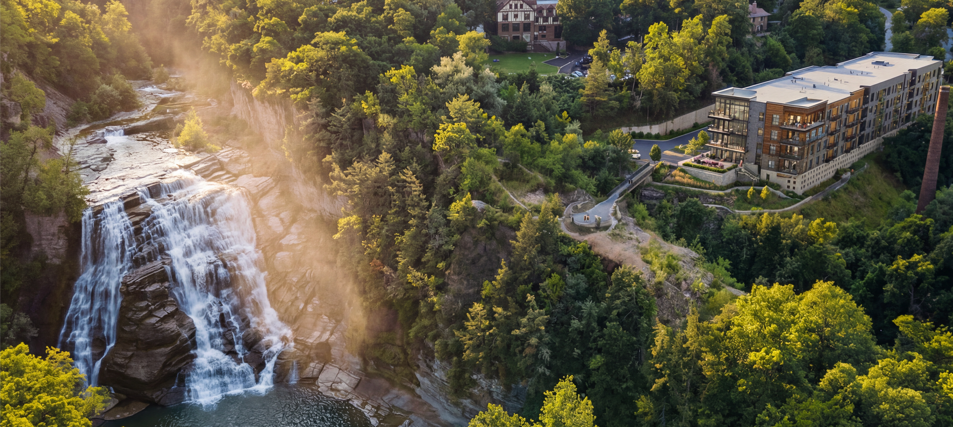 Aerial view of a waterfall cascading into a pool below, with a building perched on a cliff edge surrounded by trees.