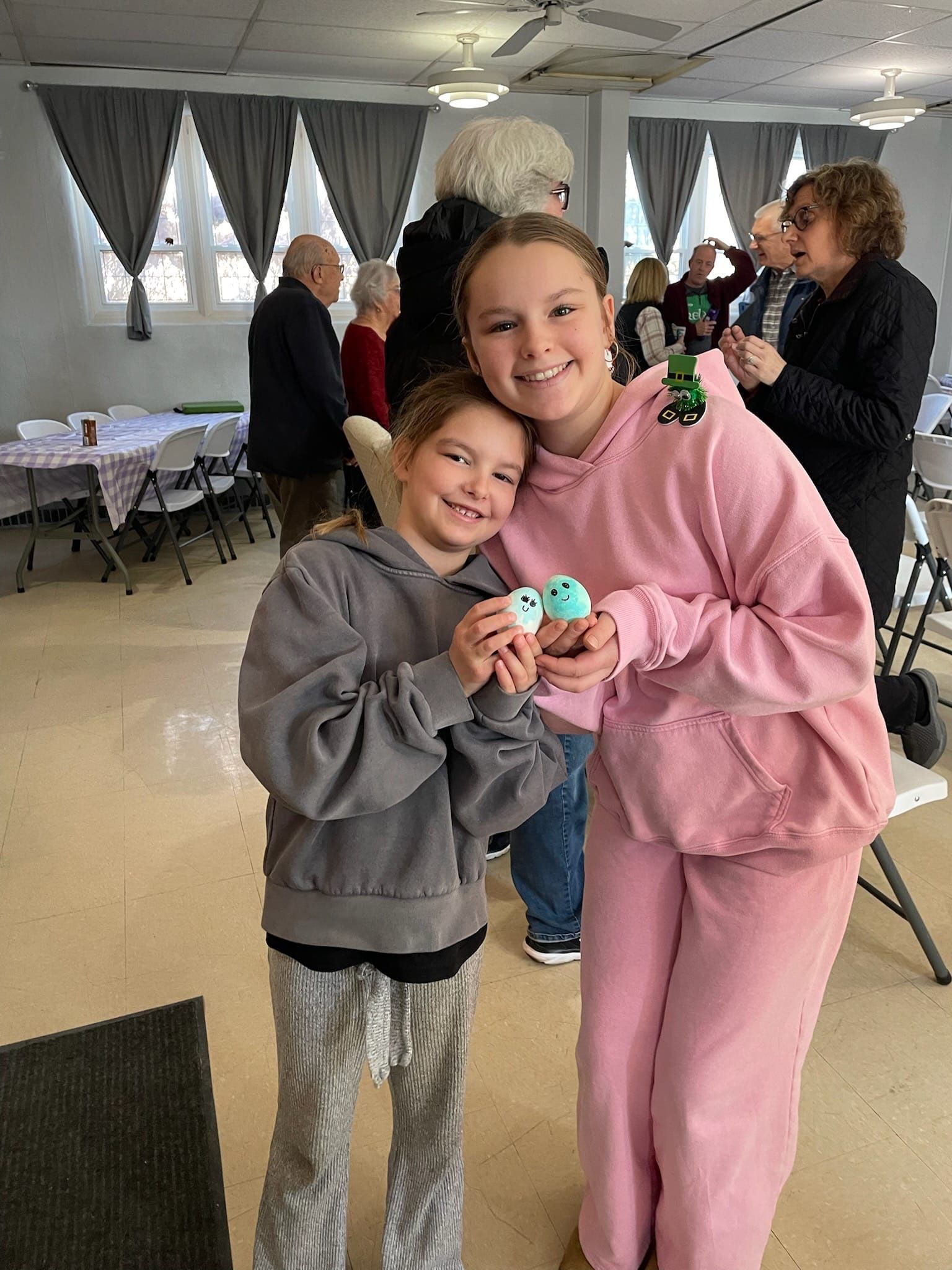 Two children smiling and holding a small toy in a decorated indoor room with people in the background.