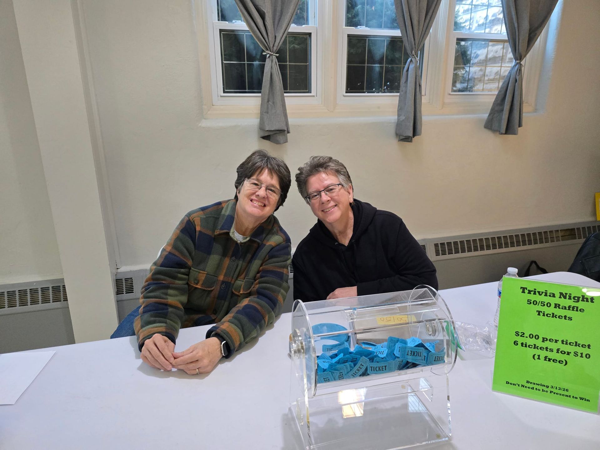 Two people sit behind a table with a clear donation box and a bright green sign in a room with windows.
