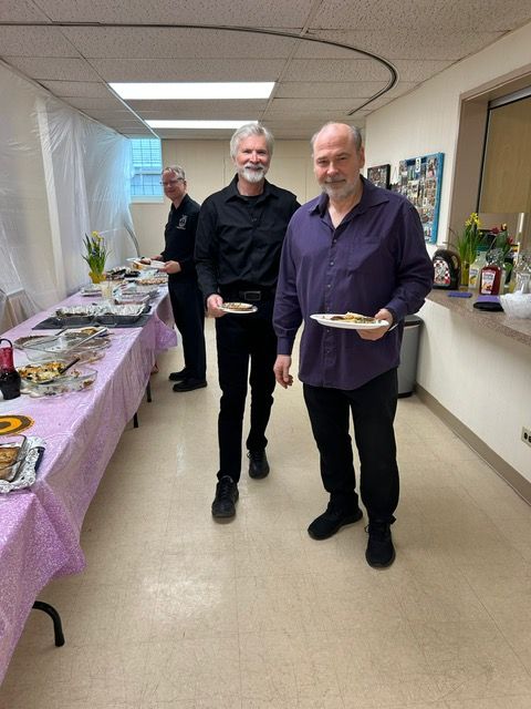 Two men holding plates at a buffet table in a hallway, with a third person in the background