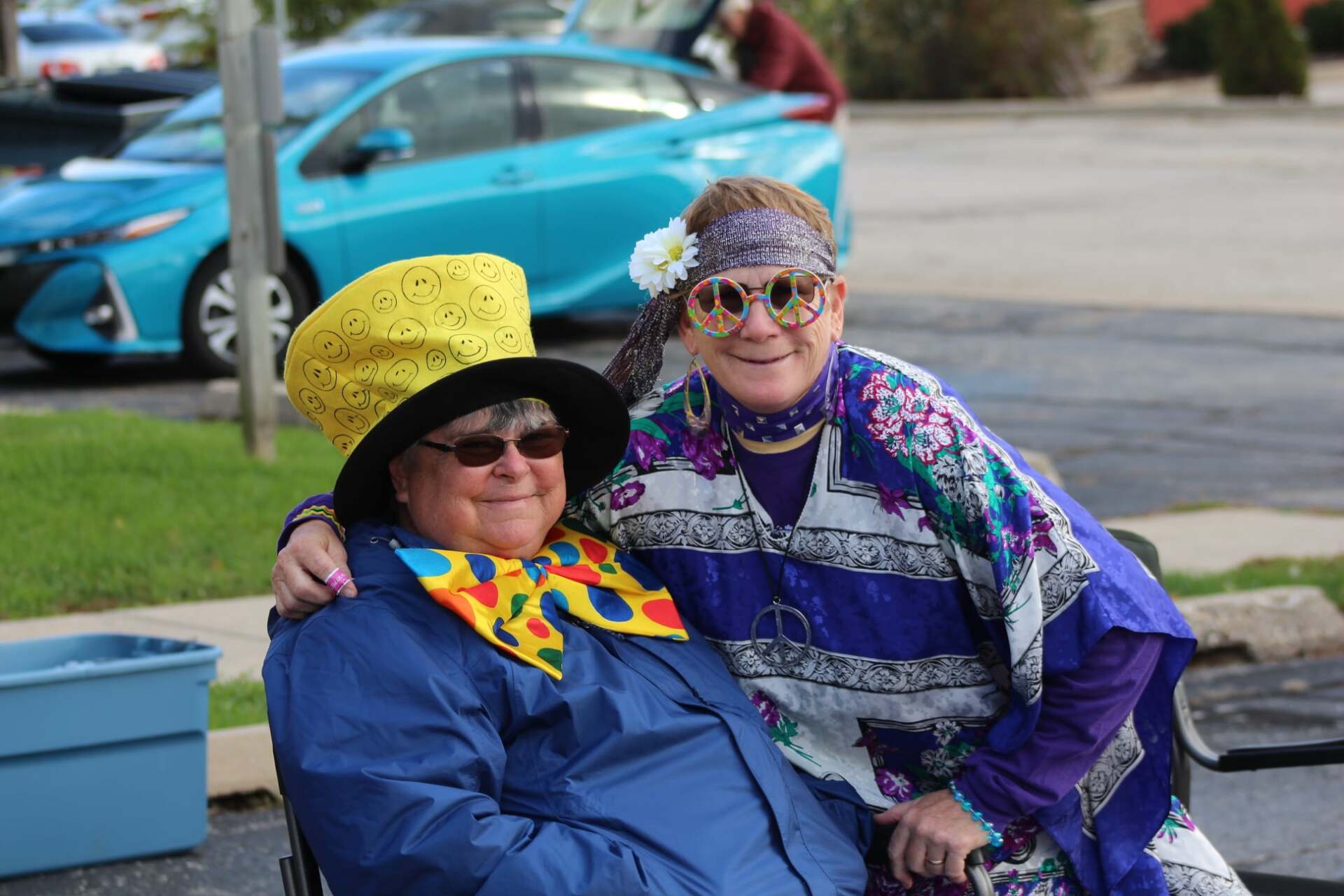 Two women are posing for a picture in front of a blue car . one of the women is wearing a clown costume.